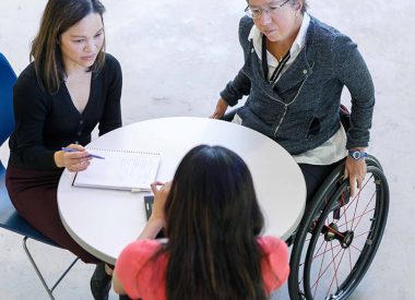 Three people, including two in wheelchairs, sit at a table discussing a document